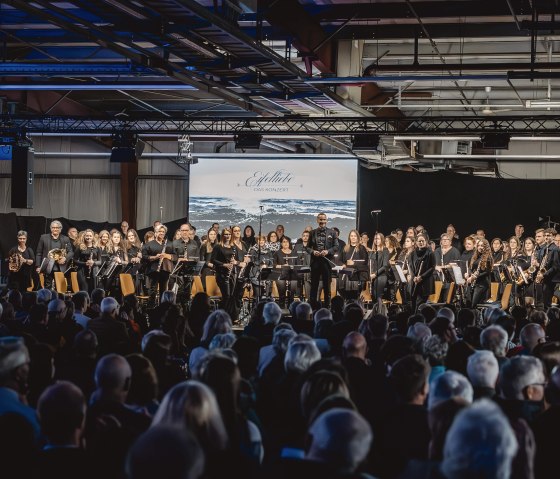 Dunkle Halle, in der sich ein großes Orchester vor seinem Publikum erhebt.