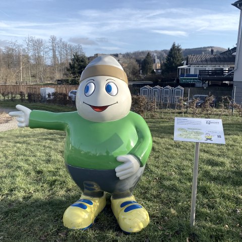 Eine Lebensgro&szlig;e Figur des Maskottchens Willi Basalt mit gr&uuml;nem Oberteil, grauer Hose, gelben Schuhen und einer M&uuml;tze auf dem Kopf. Die Figur lacht freundlich und zeigt in einen Park hinein.