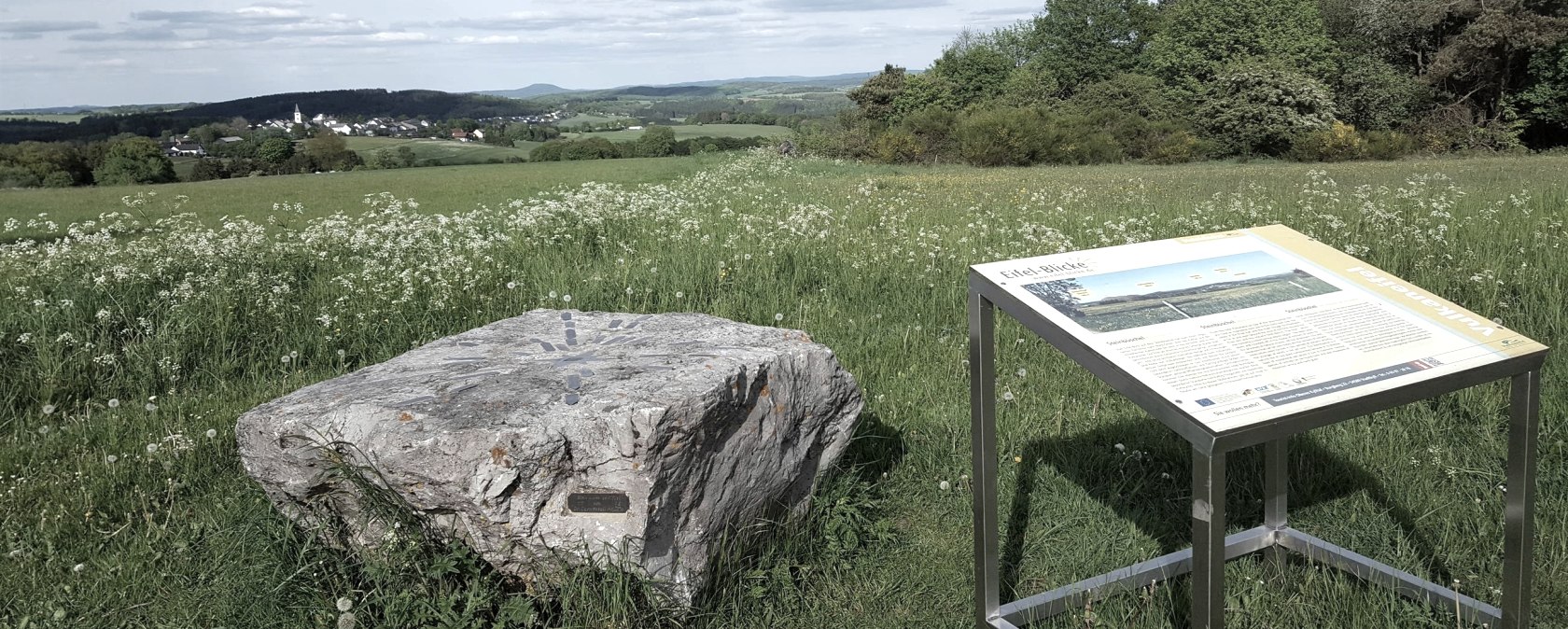 Grüne Wiese mit Felsen und Infotafel, im Hintergrund ein Dorf und Hügel unter blauem Himmel., © Touristik GmbH Gerolsteiner Land