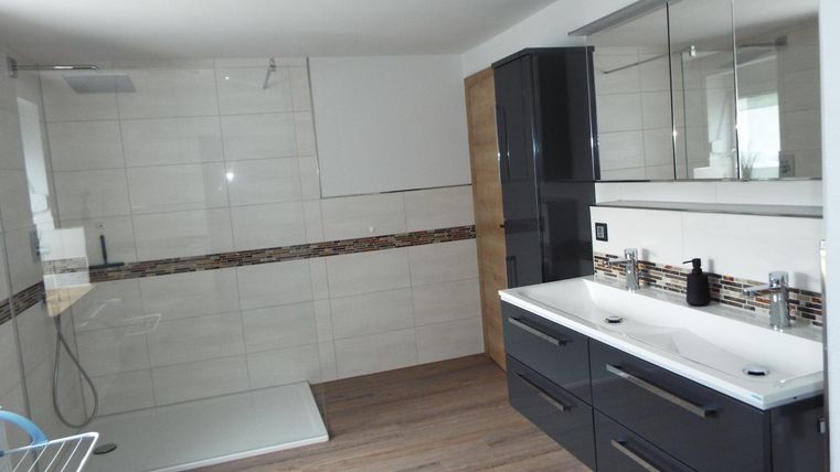 Ein modernes Badezimmer mit einer großen Dusche und einem eleganten Waschtisch. Der Boden ist mit Holzoptik gestaltet und die Wände sind mit hellen Fliesen versehen.