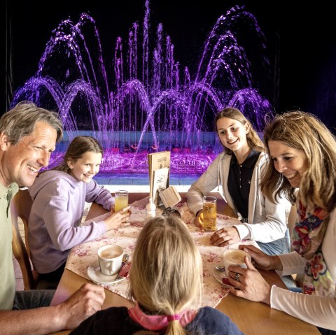 Een man en een vrouw zitten met hun drie dochters aan tafel en drinken koffie of frisdrank. Achter de familie spuit een kleurrijk waterorgel water in de lucht.