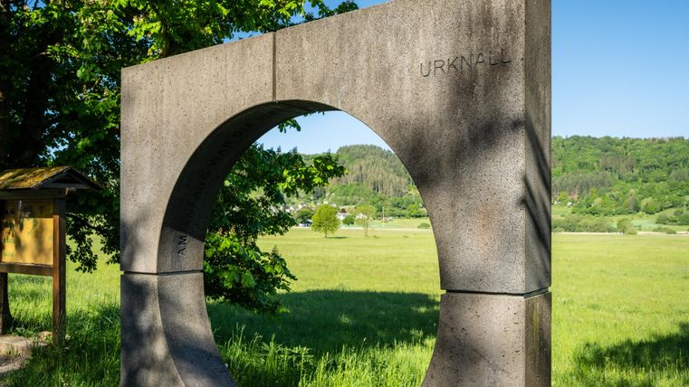 Ein kreisförmiges Steinmonument steht in einer grünen Landschaft. Im Hintergrund sind Bäume und Hügel sichtbar.