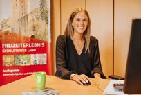Eine Frau sitzt an einem Schreibtisch und lächelt in die Kamera. Hinter ihr einige Schränke und ein Banner mit Aufdruck "Freizeirerlebnis Gerolsteiner Land" und Bild einer Burg.
