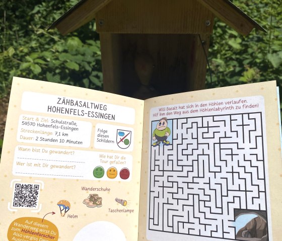 Geöffneter Wanderpass für den Zähbasaltweg in Hohenfels-Essingen mit Labyrinth und Infos zur Wanderung. Hintergrund zeigt das Stempelhäuschen im Grünen., © Touristik GmbH Gerolsteiner Land, Leonie Post