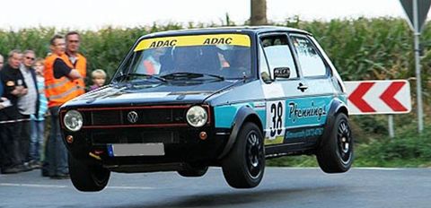 Een sportieve Volkswagen die in de lucht zweeft terwijl hij een bocht maakt. Op de achtergrond zijn toeschouwers en een groene landschap te zien.