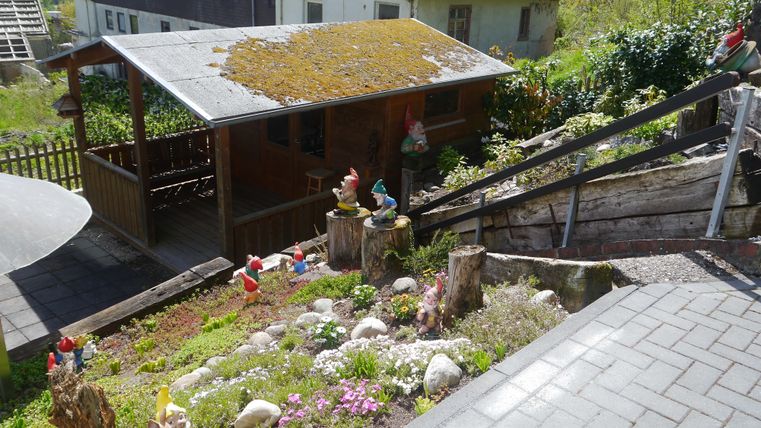 Eine rustikale Hütte mit einem begrünten Dach steht in einem Garten. Der Garten ist bunten Blumen und mehreren Gartenzwergen geschmückt.