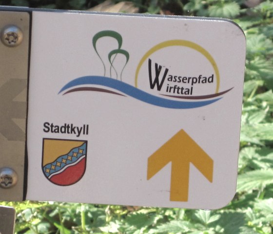 Wegwijzer naar het Wirfttal waterpad in Stadtkyll met gele pijl en wapenschild. Achtergrond van groene bladeren., &copy; Touristik GmbH Gerolsteiner Land