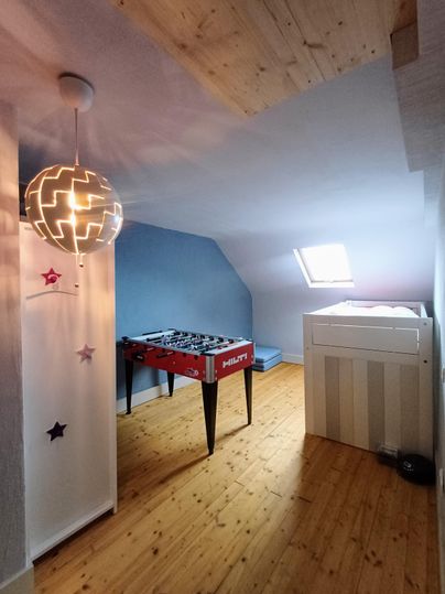 Une pièce lumineuse avec un sol en bois et des murs en pente. Au centre se trouve un baby-foot et il y a une fenêtre avec de la lumière du jour.
