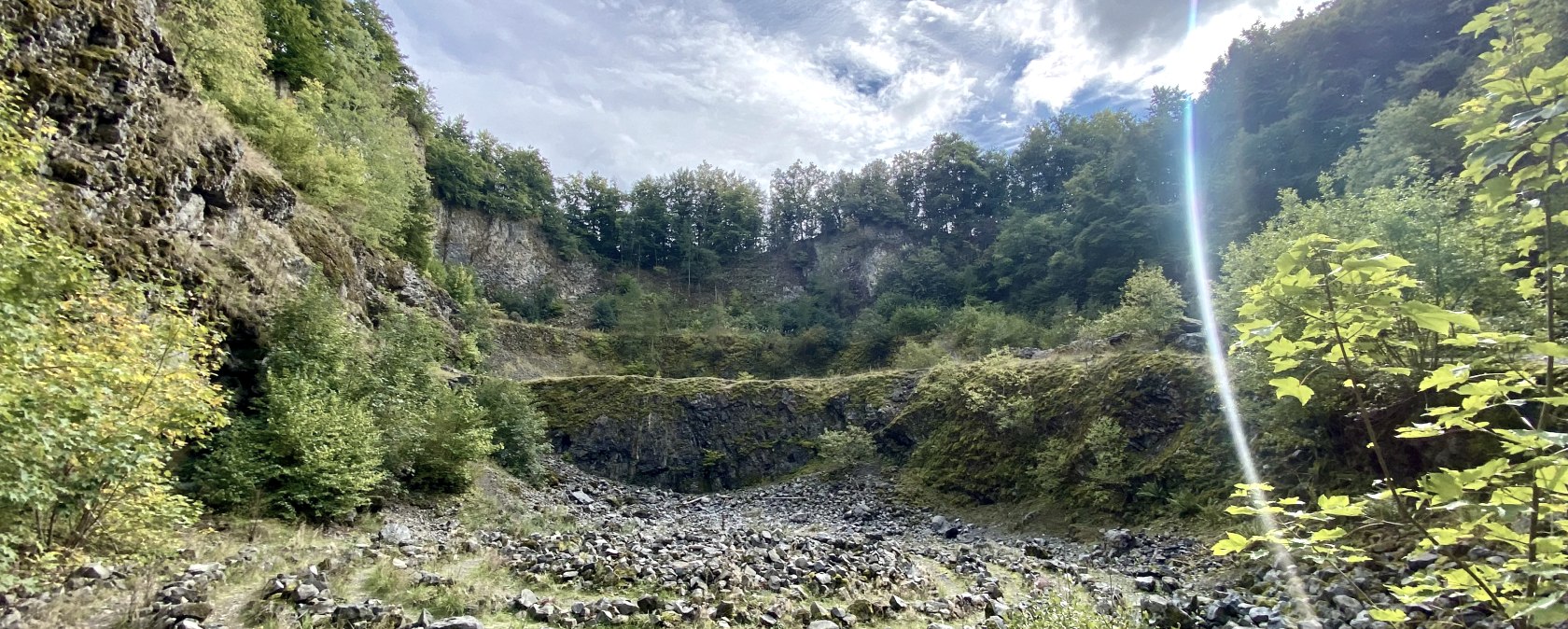 Binnenkant van de Arensberg-vulkaan met rotsachtige paden, omgeven door groene vegetatie en rotsen onder een bewolkte hemel met zonnestralen., &copy; Touristik GmbH Gerolsteiner Land