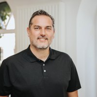 Ein Mann steht hinter einem Tresen und lächelt in die Kamera. Hinter ihr einige Fenster und Regale mit regionalen Produkten.