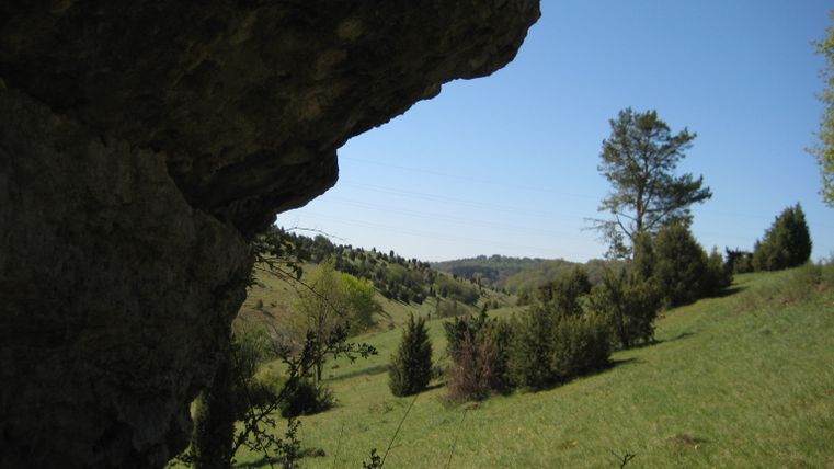 Blick durch einen Felsvorsprung auf eine grüne Hügellandschaft mit Bäumen und blauem Himmel.