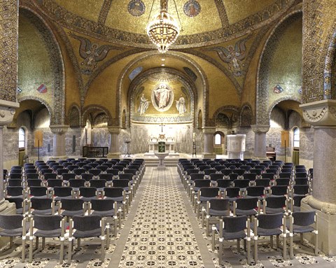 The picture shows the interior of the Church of the Redeemer with a view of the ceiling and the altar. The ceiling is decorated with golden mosaics, angels and a large chandelier. Windows let light into the magnificent church interior. Numerous chairs form rows of seats for visitors.