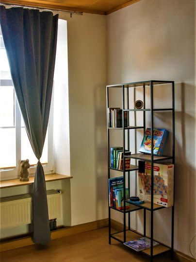Ein heller Raum mit einem Bücherregal in der Ecke. Vor einem Fenster hängt ein grauer Vorhang.