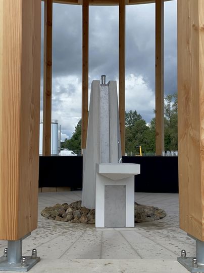 Een moderne waterpartij van beton met een gootsteen onder een open houten constructie, op de achtergrond een bewolkte lucht en bomen.