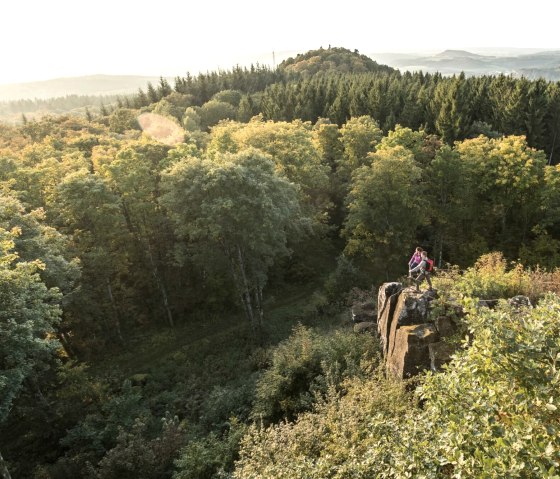 Two people are standing on a rock surrounded by a dense forest. Hills and a wide view over the landscape can be seen in the background., © Eifel Tourismus GmbH, D. Ketz