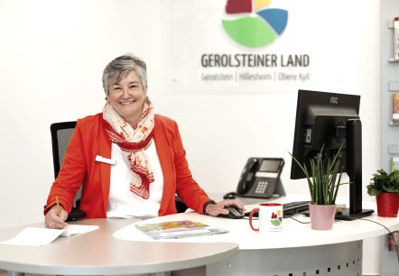 A woman sits at a desk in a bright office with a computer, telephone and keyboard in front of her and smiles at the camera. Behind her are brochure racks from the tourist information office and the logo of Touristik GmbH Gerolsteiner Land.