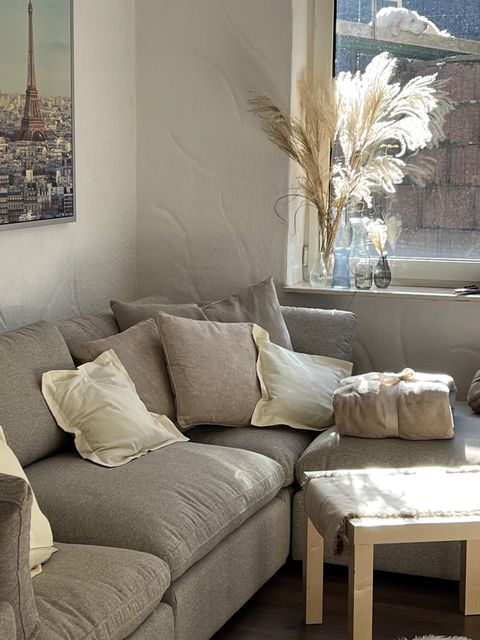 Een gezellige zithoek met een grijze bank en veel kussens. Naast het raam staat een decoratieve vaas met pampasgras.