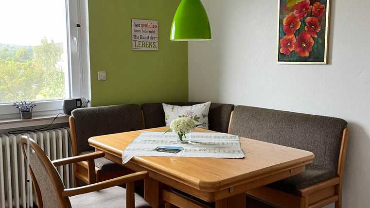 Une zone de repas confortable avec une table en bois et des meubles rembourrés beiges. Au mur, une image avec des fleurs rouges est accrochée et une lampe verte pend du plafond.