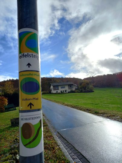 Un panneau indicateur sur un poteau montre différents sentiers de randonnée dans l'Eifel. En arrière-plan, on peut voir une maison et une pelouse verte sous un ciel nuageux.