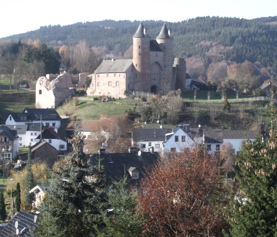 Een middeleeuws kasteel met twee torens torent uit boven een dorp in een heuvelachtig, bebost landschap., © Touristik GmbH Gerolsteiner Land