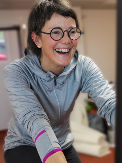 Une femme joyeuse s'entraîne sur un vélo. Elle porte un sweat à capuche gris et rit de tout cœur.