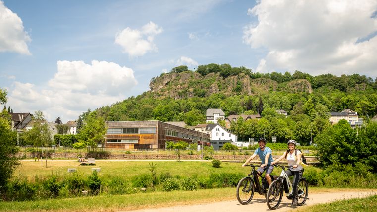 Ein Mann und eine Frau fahren auf Fahrrädern bei schönem Wetter durch den Kurpark Gerolstein mit Dolomitfelsen im Hintergrund.