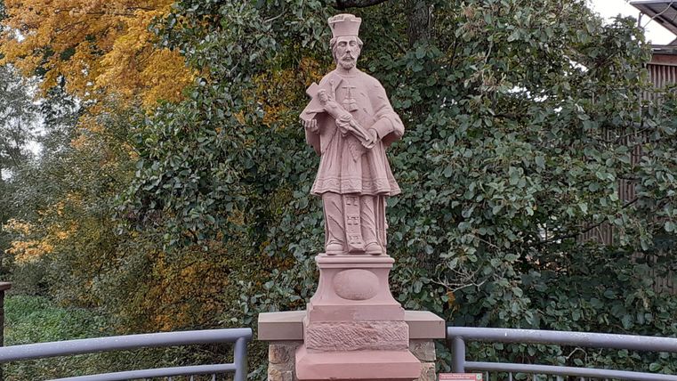 Statue des hl. Johannes von Nepomuk auf einer Brücke, umgeben von Herbstbäumen.