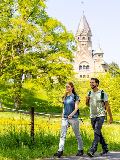 Zwei Wanderer gehen auf einem Weg, im Hintergrund eine Kirche in Mirbach, umgeben von grüner Landschaft.