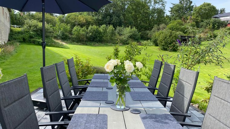 Ein schön gedeckter Tisch im Freien mit grauen Stühlen und einem Sonnenschirm. Im Mittelpunkt steht eine Vase mit weißen Rosen, umgeben von einer grünen Gartenlandschaft.