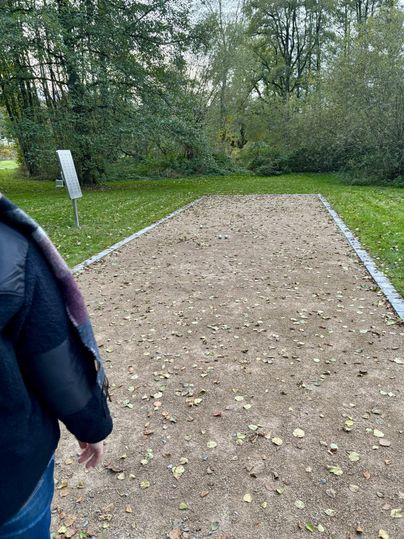 Ein Sandplatz mit vereinzelten Laubblättern in einer grünen Umgebung. Im Hintergrund sind Bäume und ein Informationsschild zu sehen.