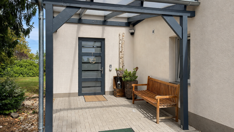 L'entrée d'une maison avec une porte d'entrée moderne et un banc en bois. Il y a des plantes décoratives et un sol pavé.