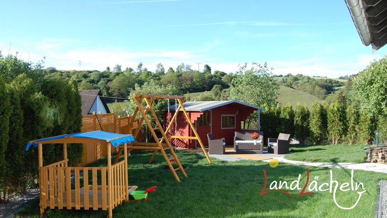 Ein schöner Garten mit Spielgeräten für Kinder und gemütlichen Sitzmöglichkeiten. Umgeben von grünen Hügeln und einer entspannten Atmosphäre.