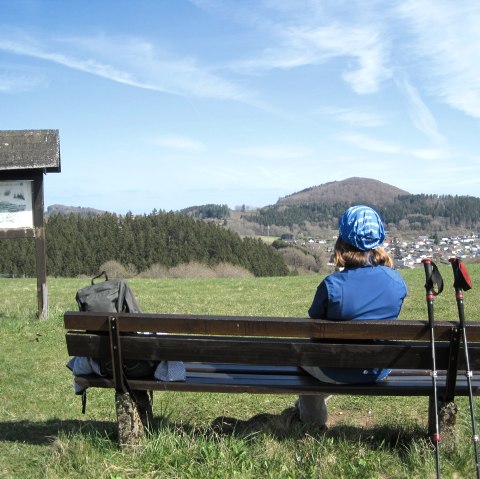 A person sits on a bench and looks at the Nerother Kopf. Hiking poles lean against the bench, a rucksack lies next to it. The sky is blue., &copy; Touristik GmbH Gerolsteiner Land, Ute Klinkhammer