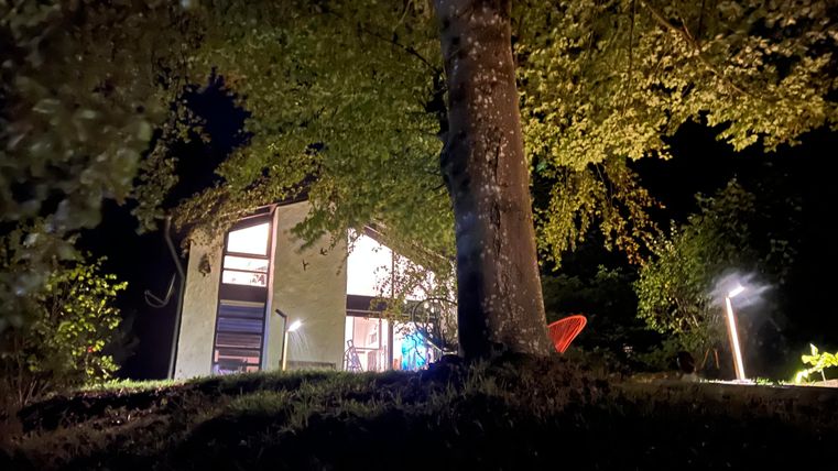 Een modern huis in de nacht, omgeven door bomen. Zacht licht verlicht het buitengedeelte en creëert een gezellige sfeer.