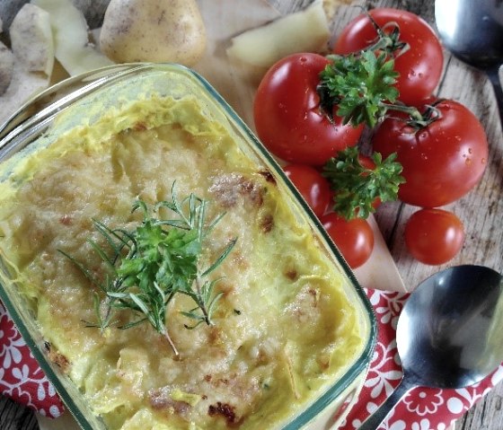 A glass baking dish with a cheese-topped potato dish in it. Next to it are tomatoes, potatoes and spoons.