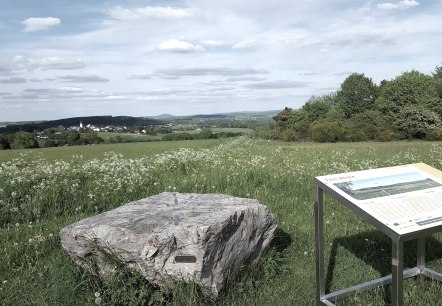Grüne Wiese mit Felsen und Infotafel, im Hintergrund ein Dorf und Hügel unter blauem Himmel., © Touristik GmbH Gerolsteiner Land