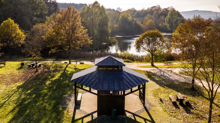 Ein Pavillon in einer herbstlichen Parklandschaft mit Bäumen und einem See im Hintergrund.