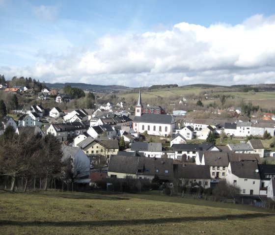 View of Stadtkyll with church in the center, surrounded by houses and hills under a blue sky with clouds., © Touristik GmbH Gerolsteiner Land