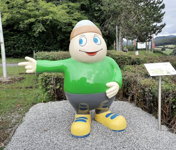 Standbeeld van mascotte Willi Basalt met groen shirt, grijze broek en gele schoenen wijst naar links. Achter hem staan bomen en een parkeerplaats.