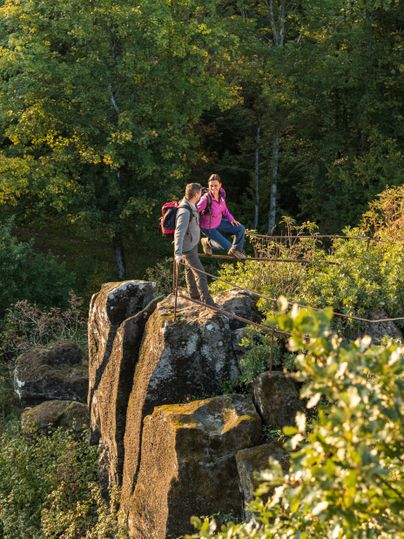 Zwei Personen stehen auf einem Felsen mit Blick auf einen Wald und Hügel im Hintergrund.