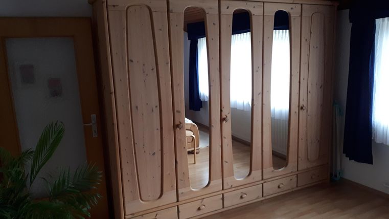 Ein großer Holzschrank mit Spiegeln und mehreren Türen steht in einem Raum. Die Fenster lassen viel Licht hereinfällen und die Möbel sind schlicht.