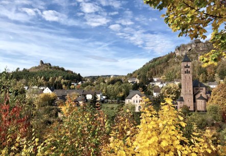 View of several residential buildings and a large church looming ahead. All around are meadows and trees in an autumnal atmosphere. Rocks rise up above the town.