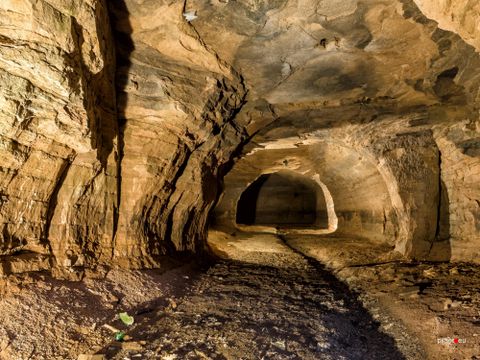 Ein beleuchteter Tunnel mit steinigen Wänden und gewölbter Decke. Der Boden ist uneben und mit Schutt bedeckt. Im Hintergrund ist eine Öffnung sichtbar.
