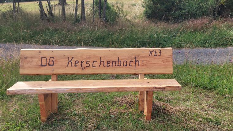 Holzbank mit der Aufschrift 'OG Kerschenbach' auf einer Wiese.