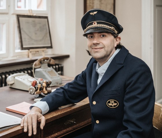 Mann in historischer Schaffnerkleidung sitzt an einem Schreibtisch, auf dem eine Schreibmaschine, Stempel und ein antikes Telefon stehen und schaut in die Kamera.