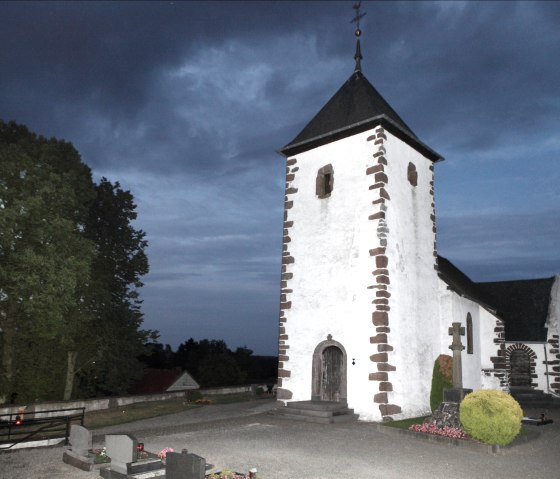 Berndorf fortified church at dusk, surrounded by trees and a cemetery. The sky is dark blue, the church has white walls with dark stones., © Regnery