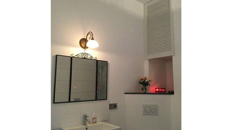 Ein modernes Bad mit einem weißem Waschbecken und einem großen Spiegel. An der Wand ist eine elegante Lampe und ein kleines Blumenarrangement zu sehen.