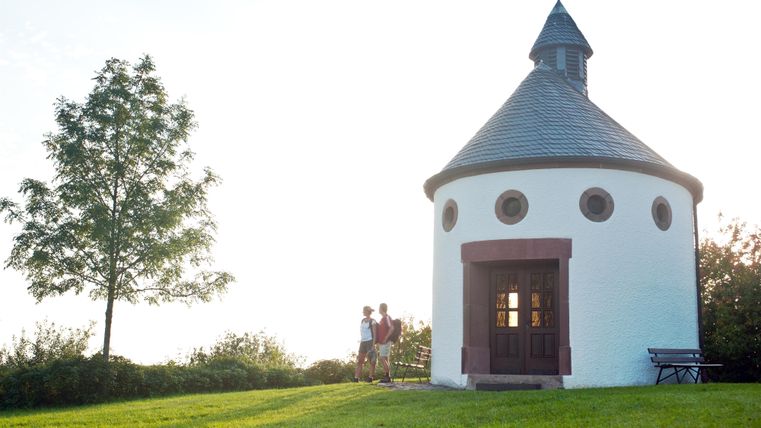 Eine kleine, runde Kapelle steht in einer grünen Wiese. Zwei Personen stehen davor und genießen die Aussicht.