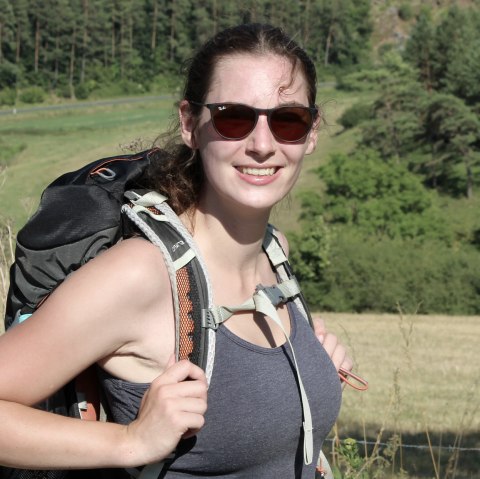 Eine Frau auf einer Wiese mit Wanderrucksack auf dem Rücken und Sonnenbrille schaut lachend zur Kamera.