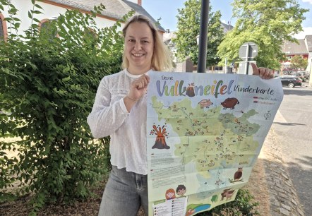 Een vrouw staat op een plein voor een heg en houdt een grote kaart met de titel &ldquo;Vulkaneifel-kinderkaart&rdquo; omhoog.